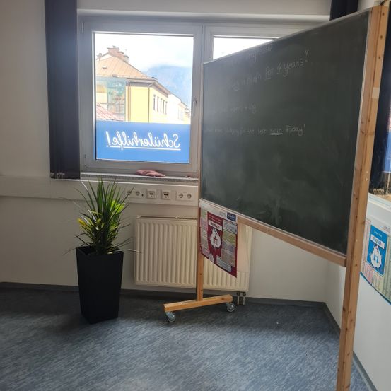 Ein leeres Klassenzimmer mit einer großen Tafel mit Schrift, einem Topfpflanzen und einem Fenster mit einem blauen Schild.
