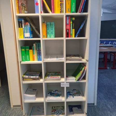 Ein Bücherregal mit Büchern zu verschiedenen Themen, einschließlich Mathematik, Englisch und Latein, steht in einem Raum mit einem roten Schreibtisch und einer Tafel. Das Regal hat Fächer für Bücher, und das Preisschild unten links lautet 'Hefte a 2e'.