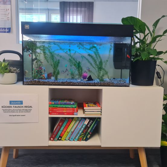 Ein weißer Schrank beherbergt ein Aquarium mit Fischen und Pflanzen. Ein Bücherregal darunter hat bunte Bücher. Ein Schild steht für 'Bücher-Tausch-Regal' und eine Werbung ist an der Wand.