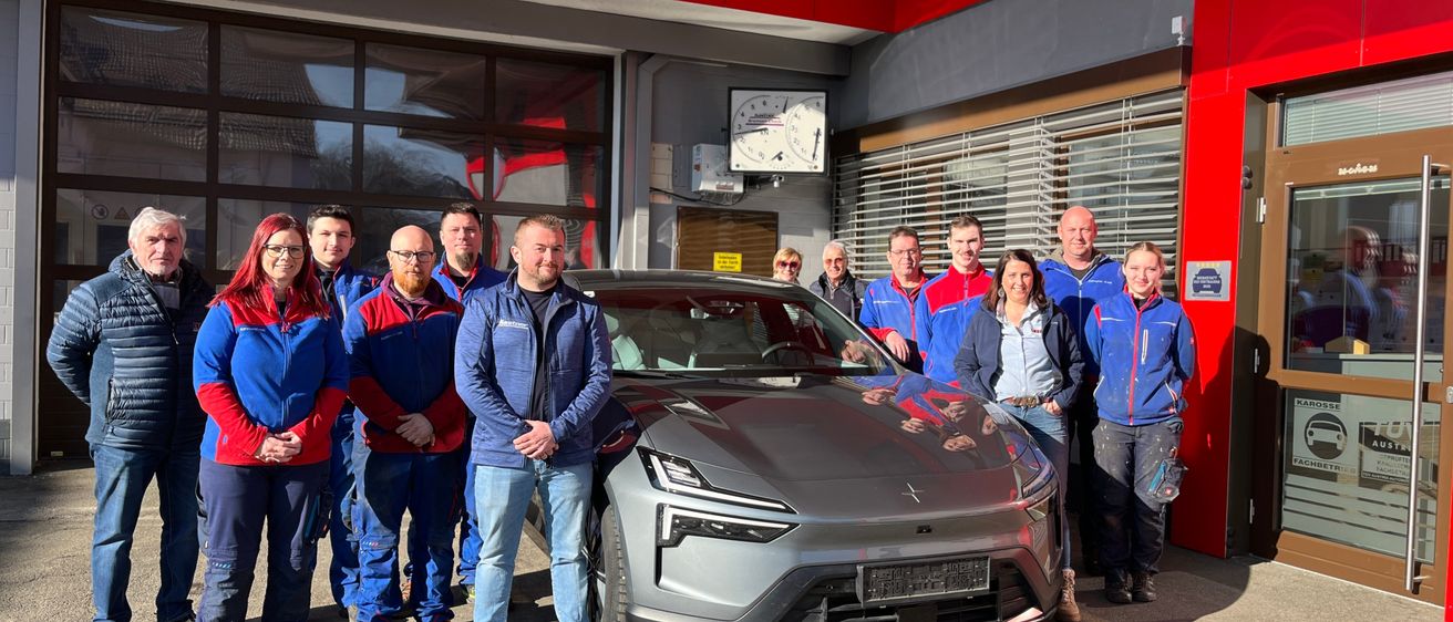 Eine Gruppe von Menschen in passenden blauen und roten Jacken steht vor einem schlanken, grauen Elektroauto vor einem roten Gebäude.