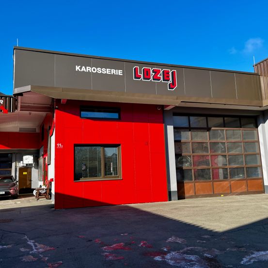 Ein rotes Gebäude mit dem Schild 'LOZEJ KAROSSERIE' über dem Eingang. Ein Auto ist in der Garage geparkt.