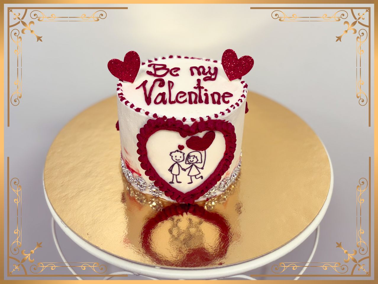 Ein weißer Kuchen mit roten Herzen oben drauf, die 'Sei mein Valentin' sagen. Es hat ein herzförmiges Design und ein Paar, das sich an den Händen hält. Es steht auf einem goldenen Teller.