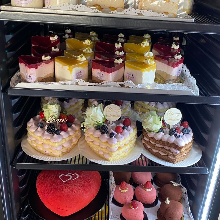 Eine Vitrine voller verschiedener Desserts. Drei herzförmige Kuchen mit unterschiedlichen Toppings sind prominent ausgestellt. Andere Desserts umfassen Mini-Kuchen und Cupcakes, einige mit herzförmigen Etiketten.
