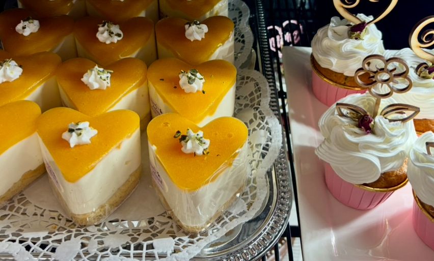 Mehrere herzförmige Käsekuchen sind auf einem Tablett ausgestellt, gekrönt mit Schlagsahne und einer dekorativen Blume. Daneben stehen zwei Cupcakes mit rosa Glasur und weißer Glasur auf einem weißen Teller.