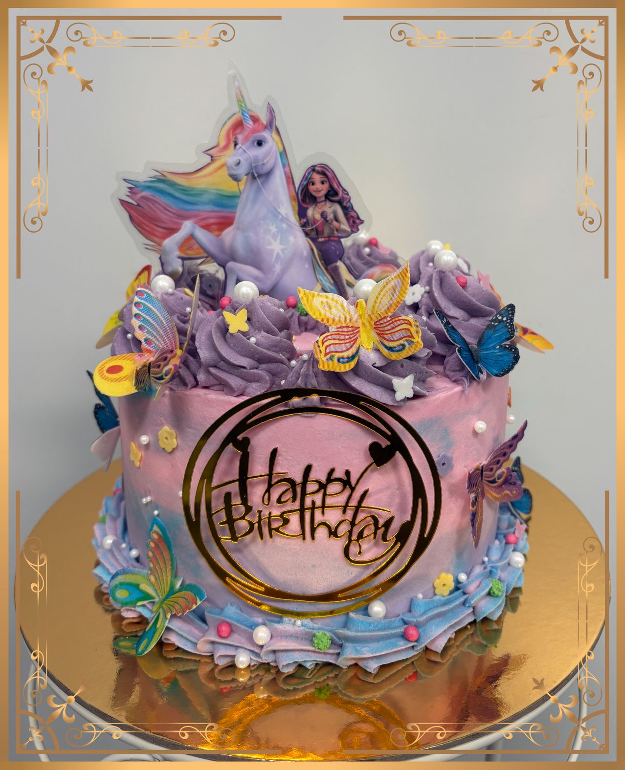 A birthday cake decorated with a unicorn, mermaid, and butterflies. The cake is purple and pink with a gold circle saying 'Happy Birthday'. It is on a golden plate with a white background.