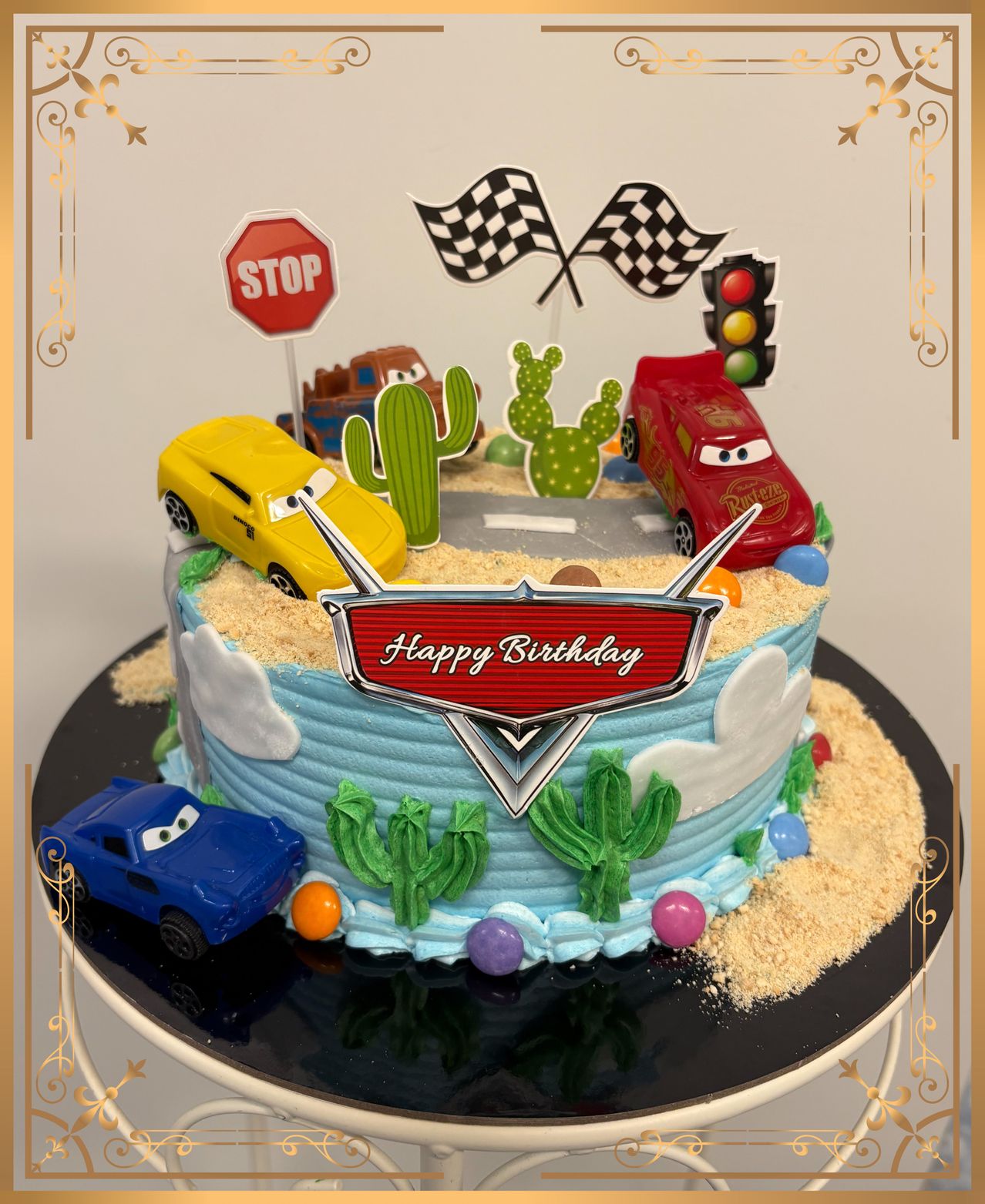 A birthday cake with a blue base and racing cars, a stop sign, and cacti. It says 'Happy Birthday' in red.