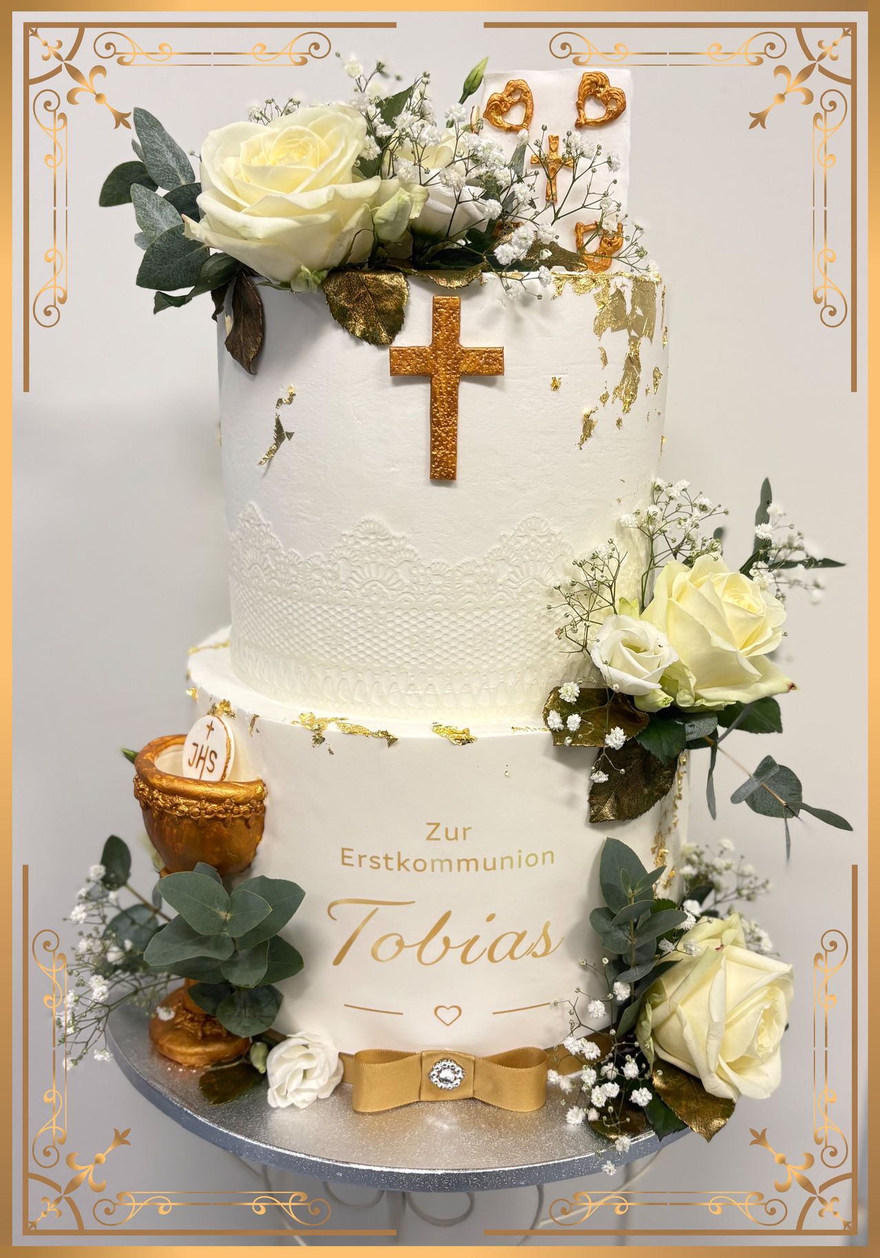 Ein dreistöckiger Kuchen mit goldenem Kreuz, Blumen und dem Namen Tobias in Gold. Verziert mit Goldfolie, weißen Rosen und grünen Blättern.