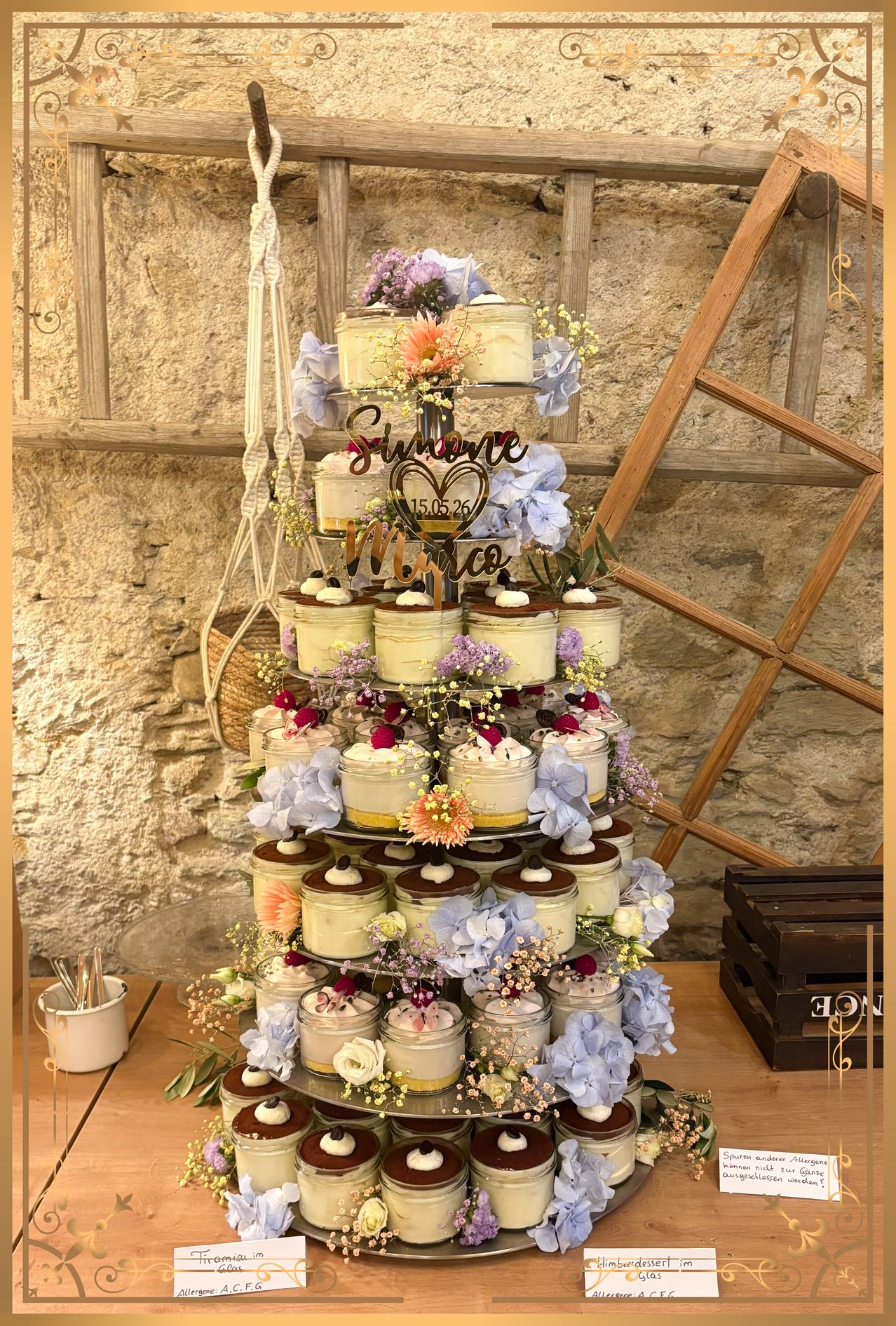 Ein mehrschichtiger Kuchenständer mit Dessertgläsern, dekoriert mit Blumen und personalisierten Namensschildern, in einer rustikalen Umgebung aufgestellt.