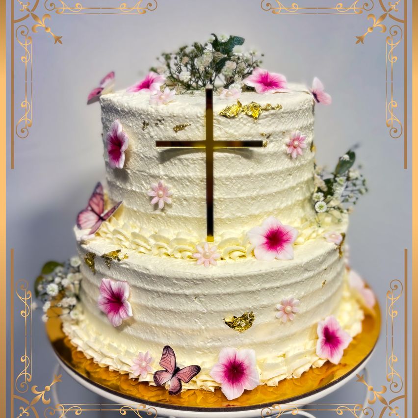 Ein dreistöckiger Kuchen mit weißer Glasur und rosa Blumen, gekrönt von einem goldenen Kreuz und Schmetterlingen. Der Kuchen hat einen goldenen Rand und ist mit kleinen Blumen verziert.
