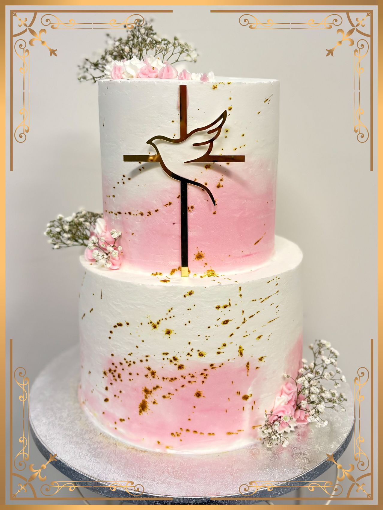 A three-tiered cake with a gold cross and dove design, decorated with pink icing, gold sprinkles, and small flowers.