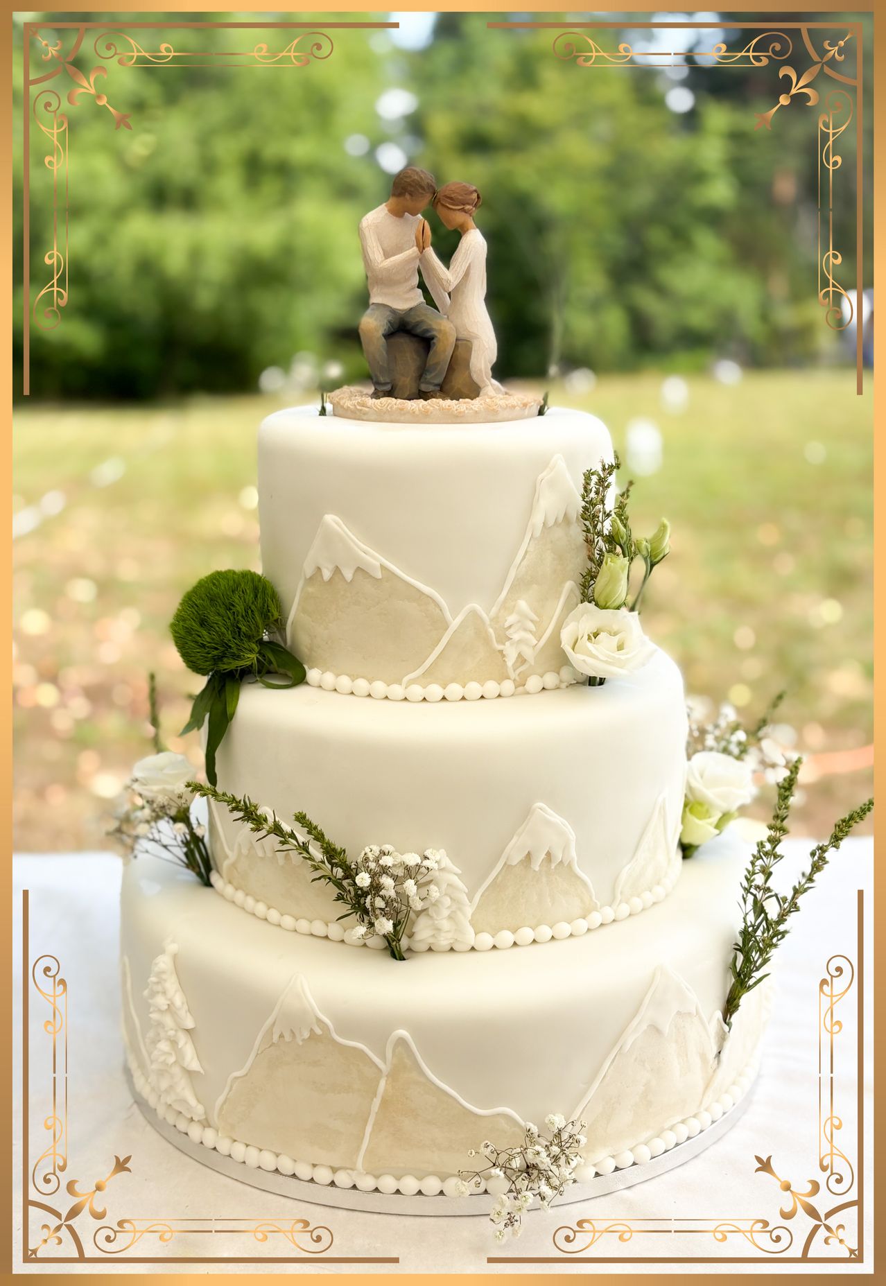 A three-tiered wedding cake with a mountain theme, featuring a couple figurine on top, surrounded by white flowers and greenery.