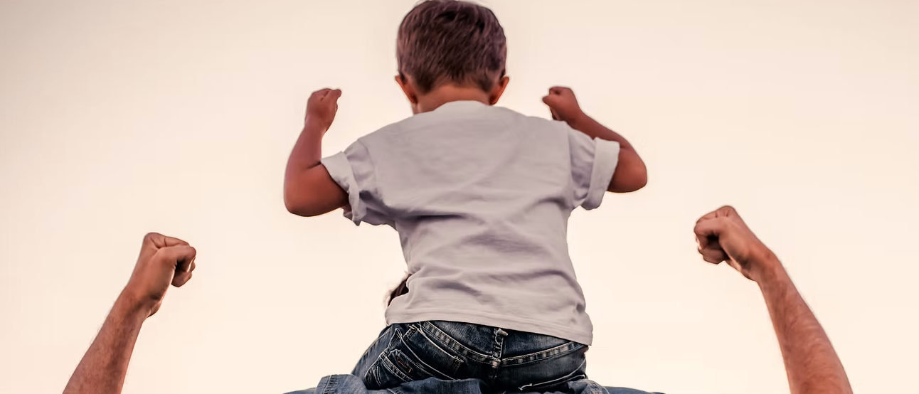 Ein junger Junge sitzt auf dem Rücken eines Erwachsenen, beide tragen Jeans. Der Junge trägt ein weißes T-Shirt und hat die Arme erhoben.