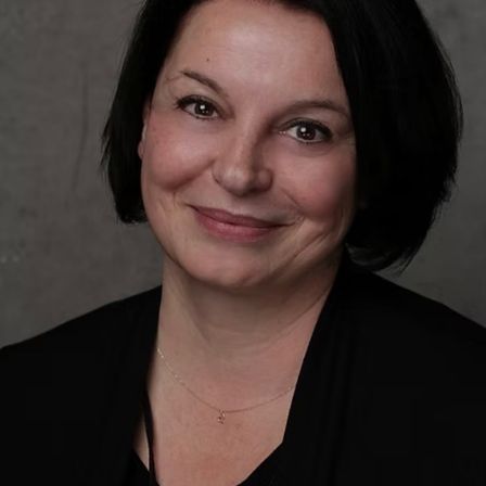 Eine Frau mit kurzen schwarzen Haaren lächelt und trägt ein schwarzes Oberteil und eine Halskette. Sie hat einen Muttermal auf ihrer rechten Wange.