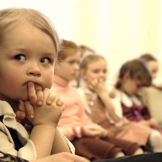 Ein kleines Kind mit den Händen am Mund sitzt aufmerksam in einer Reihe mit anderen Kindern, alle in pinker und brauner Kleidung.