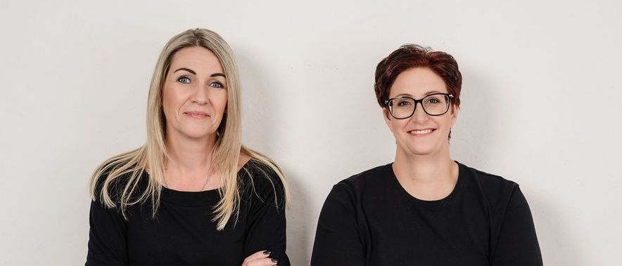 Zwei Frauen stehen nebeneinander mit verschränkten Armen. Beide tragen schwarze Langarmshirts und Jeans. Die Frau auf der linken Seite hat blonde Haare und ein Tattoo am linken Arm. Die Frau auf der rechten Seite trägt eine Brille. Sie lächeln.