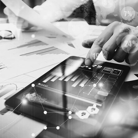 Eine Hand zeigt auf ein Tablet mit einem Diagramm und Zahlen, auf einem Schreibtisch mit Papieren und einer Karte. Eine verschwommene Person sitzt in der Nähe.