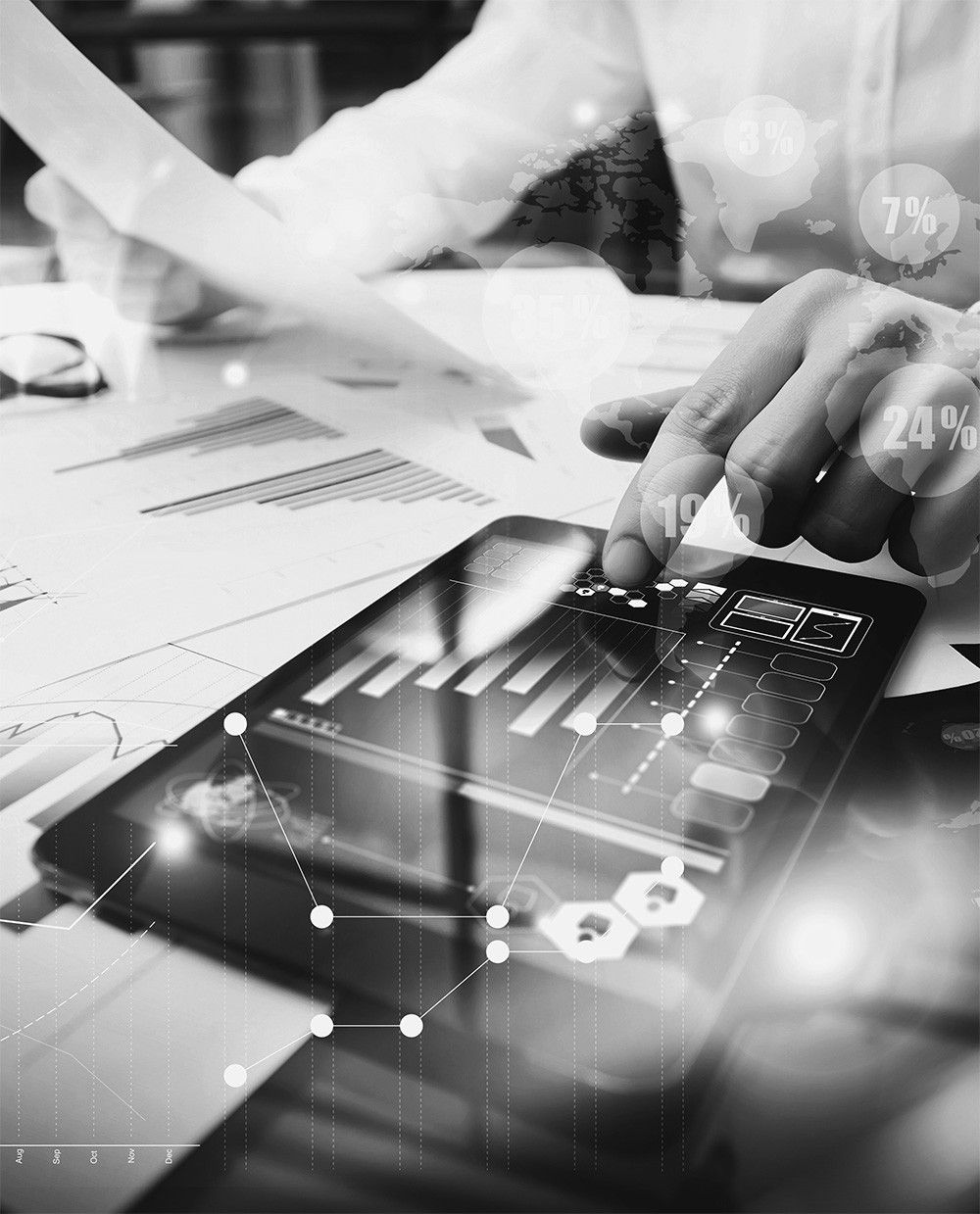 Eine Hand zeigt auf ein Tablet mit einem Diagramm und Zahlen, auf einem Schreibtisch mit Papieren und einer Karte. Eine verschwommene Person sitzt in der Nähe.