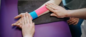 Eine Hand mit einem bunten Verband am Unterarm ruht auf einer violetten Oberfläche und wird von einer anderen Person gehalten.