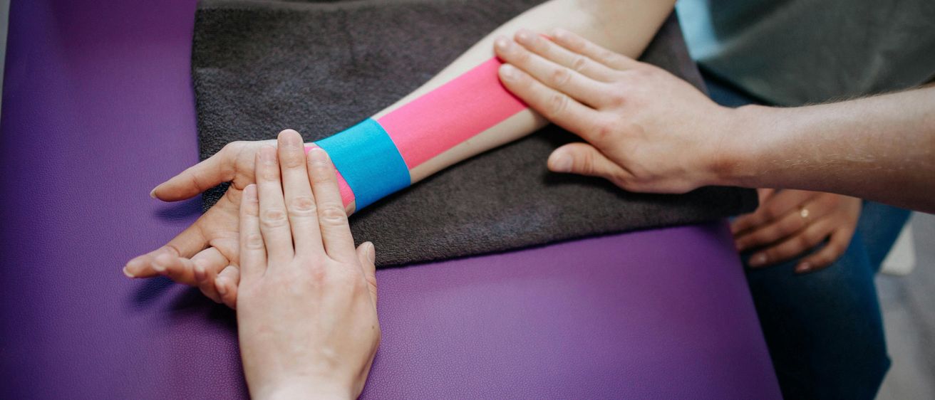 Eine Hand mit einem bunten Verband am Unterarm ruht auf einer violetten Oberfläche und wird von einer anderen Person gehalten.