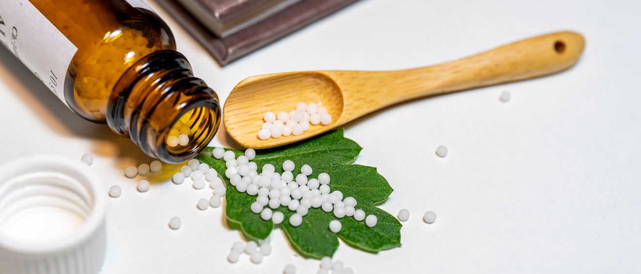 Eine Flasche mit homöopathischen Pellets und ein hölzerner Löffel liegen auf einer weißen Oberfläche, mit einem Blatt in der Nähe und einem braunen Buch im Hintergrund.
