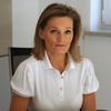 Eine Frau mit blonden Haaren sitzt an einem Schreibtisch und trägt ein weißes Polo-Shirt mit einem Logo auf der linken Brust. Sie sieht ernst und konzentriert aus, möglicherweise arbeitet oder nimmt an einer Besprechung teil.