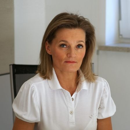 Eine Frau mit blonden Haaren sitzt an einem Schreibtisch und hält einen grünen Stift. Sie trägt ein weißes Shirt mit einem Logo auf der linken Brust.