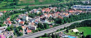 Luftaufnahme einer Stadt mit vielen Gebäuden, roten Dächern und einer Autobahn. Grüne Bäume und Gras umgeben das Gebiet. Ein roter Zug fährt auf der Schiene.