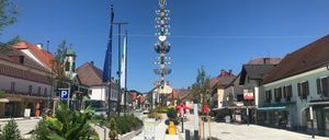 Eine Straße mit bunten Blumen und einem hohen dekorativen Turm in der Mitte. Gebäude säumen die Seiten, und Flaggen sind gehisst. Unter einem klaren blauen Himmel.