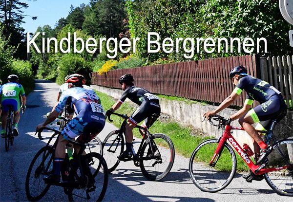 Eine Gruppe von Radfahrern in Trikots mit Nummern konkurriert auf einer asphaltierten Straße. Die Radfahrer sind von einem Zaun und Grün umgeben. Der Text Kindberger Bergrennen ist oben im Bild angezeigt.