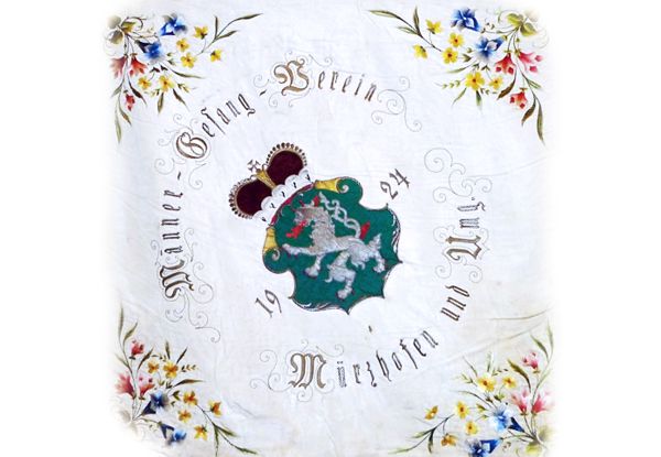 Ein antikes Taschentuch mit einem Blumenrand und einem zentralen Emblem. Das Emblem zeigt einen Löwen, eine Krone und die Zahl 24. Das Taschentuch trägt die Aufschrift 'Männer-Gesang-Verein' und 'Mit Uns'. Das Datum 19 ist ebenfalls sichtbar.