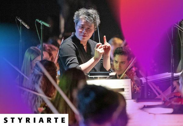 Ein Orchesterdirigent leitet eine Aufführung und hält einen Taktstock in einem hell beleuchteten Konzertsaal.