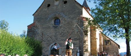Zwei Personen treten auf einer Bühne vor einer alten Kirche auf. Einer von ihnen hält einen Stock. Das Publikum schaut zu.
