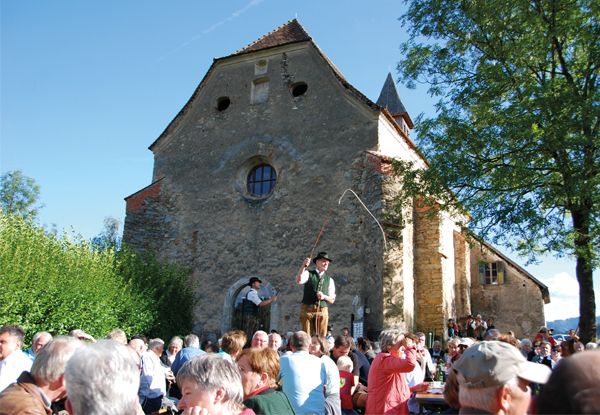 Zwei Personen treten auf einer Bühne vor einer alten Kirche auf. Einer von ihnen hält einen Stock. Das Publikum schaut zu.