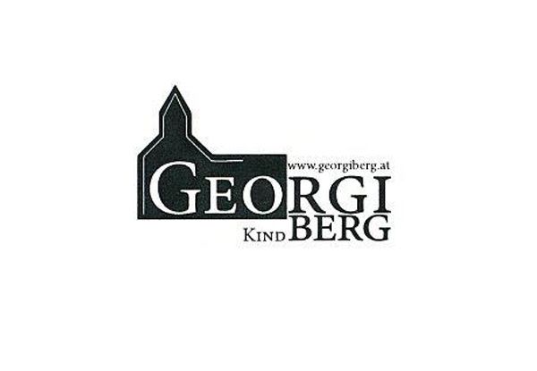 Das Georgi Kindberg Logo zeigt ein schwarz-weißes Design mit dem Text 'Georgi Kindberg' über einer kirchenähnlichen Struktur. Die Website-Adresse 'www.georgiberg.at' befindet sich unter dem Text.