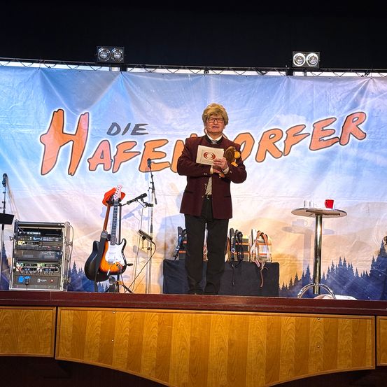 Ein Mann steht auf einer Bühne mit einem Banner, das 'Die Hafendorfer' liest. Er hält eine Auszeichnung und steht neben einem Mikrofon und einer Gitarre. Ausrüstung ist auf der Bühne und eine Tasse steht auf einem Tisch.