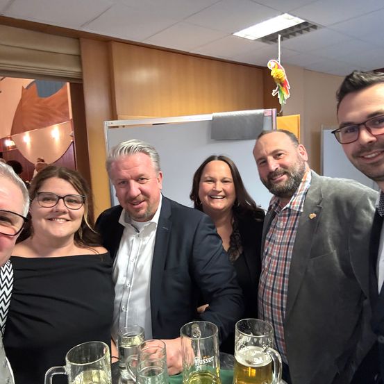Fünf Erwachsene in Businesskleidung posieren für ein Foto in einem Besprechungsraum. Biergläser auf dem Tisch.