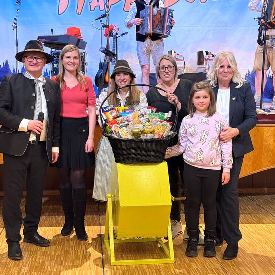 Eine Gruppe von Menschen posiert für ein Foto auf einer Bühne mit einem Korb voller Snacks. Ein Mann spielt im Hintergrund Akkordeon.