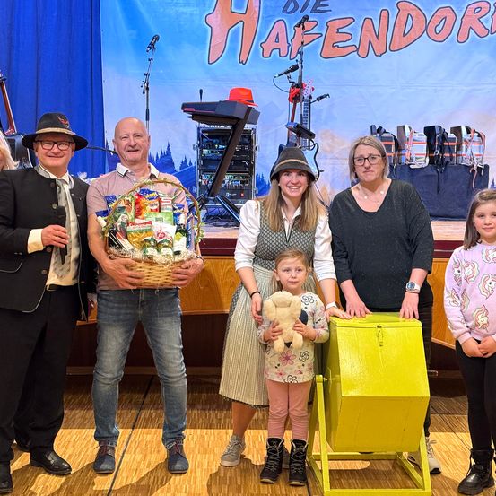 Eine Familie steht auf der Bühne und hält einen Geschenkkorb, während ein Kind einen Plüschtier hält. Eine Frau und ein Mädchen stehen vor einem gelben Container. Dahinter stehen ein Mann und eine Frau, und ein Banner mit dem Text 'DIE HAFENDOR' ist zu sehen.