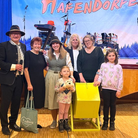 Eine Gruppe von Menschen posiert für ein Foto auf einer Bühne mit einem Banner, das 'Hafenendorf' lautet. Sie lächeln und halten eine gelbe Box. Dahinter steht ein hölzernes Podium mit einem Mikrofon.