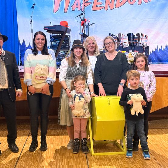 Eine Gruppe von Erwachsenen und Kindern steht auf einer Bühne mit einer gelben Kiste und einem Banner im Hintergrund. Ein Kind hält ein Mikrofon, während andere Plüschtiere halten.