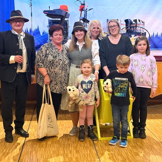 Eine Gruppe von Erwachsenen und Kindern steht lächelnd für ein Foto auf einer Bühne. Eine Frau hält ein Plüschtier und ein Mädchen ein Teddybär. Im Hintergrund sind Musikinstrumente und eine malerische Kulisse zu sehen.