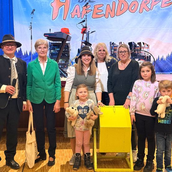 Eine Gruppe von Menschen, einschließlich Erwachsener und Kindern, posiert für ein Foto. Eine Frau in der Mitte hält ein Kuscheltier. Ein gelber Kasten steht vor ihnen. Der Hintergrund hat ein Schild mit dem Text 'Hafendorfe'.