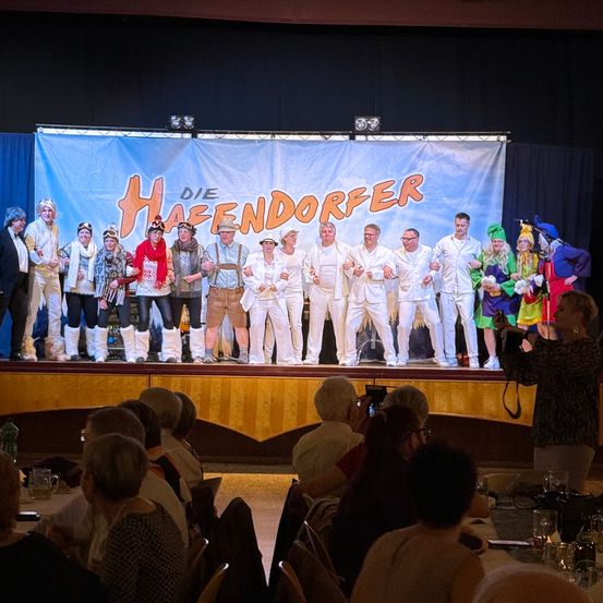 Eine Gruppe von Menschen in Kostümen steht auf der Bühne mit einem Banner, das Die Haendorfer liest. Ein Publikum beobachtet sie von vorne.