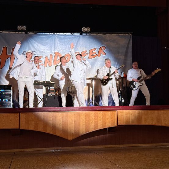 Eine Band tritt auf der Bühne auf, mit einem Banner hinter ihnen, das 'HENDER' sagt. Sie spielen Gitarren, Trommeln und eine Tastatur.