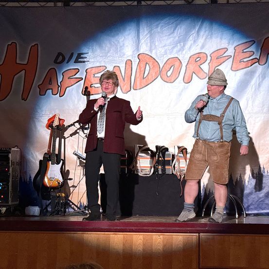 Zwei Männer stehen auf der Bühne mit Gitarren und einem Banner mit der Aufschrift 'Die Hafendorfer'. Einer gibt Daumen hoch, der andere trägt eine Mütze und eine suspender. Musikinstrumente stehen hinter ihnen.