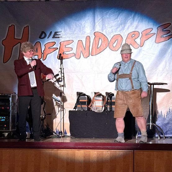 Zwei Männer stehen auf der Bühne und führen auf, einer hält ein Mikrofon und der andere singt. Sie stehen im Rampenlicht und hinter ihnen ist eine Kulisse mit 'Die Hafendorfe' in fetten Buchstaben.