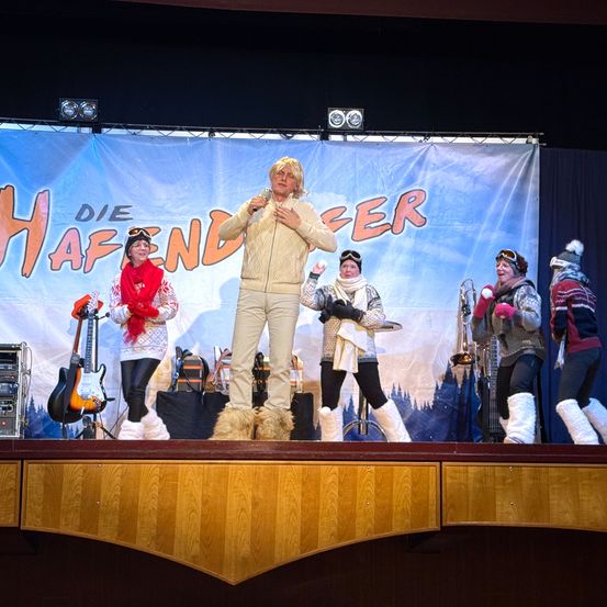 Eine Gruppe von Künstlern steht auf der Bühne unter einem Banner mit der Aufschrift Die Hafendeer. Ein Mann spricht in ein Mikrofon. Die anderen halten Musikinstrumente.