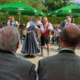Eine Gruppe von Menschen tanzt in traditioneller bayerischer Kleidung unter grünen Regenschirmen. Zuschauer sitzen um sie herum. Der Hintergrund zeigt Grün und einen klaren Himmel.