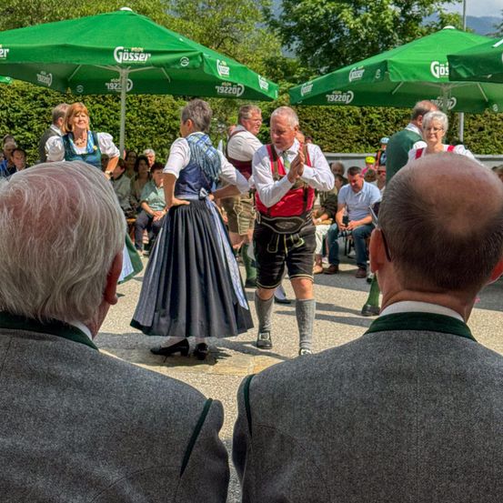 Eine Gruppe von Menschen tanzt in traditioneller bayerischer Kleidung unter grünen Regenschirmen. Zuschauer sitzen um sie herum. Der Hintergrund zeigt Grün und einen klaren Himmel.