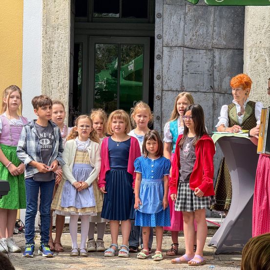 Eine Gruppe von Kindern und einer Frau steht vor einem Gebäude. Die Kinder stehen in einer Reihe, und einige tragen Turnschuhe. Die Frau steht hinter einem Tisch. Einige Kinder tragen Kleider, während andere Jacken tragen.